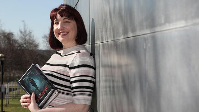 Stormi Lewis leans against a wall and holds a stack of books. The book on top is “The Protector,” written by her. 