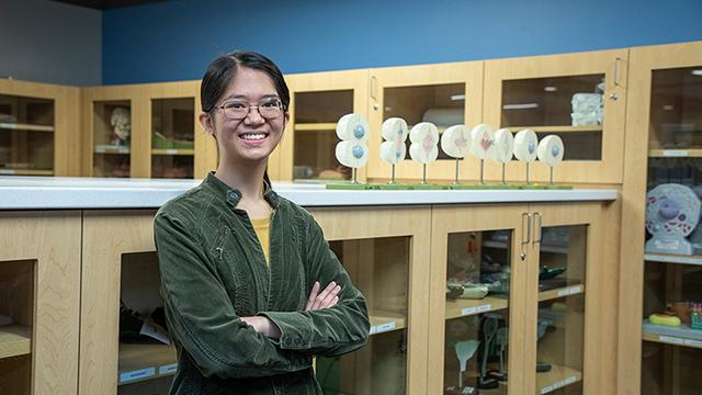 Biology tutor Kaitlyn Sy stands in a science lab room with cabinets containing scientific teaching tools around her. 在她的右边是一个展示细胞如何分裂的显示器.  